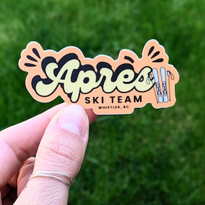 May include: A peach-coloured sticker with the word "Apres" in black and yellow lettering. The sticker also says "Ski Team Whistler, BC" and features a cartoon image of skis and ski poles.