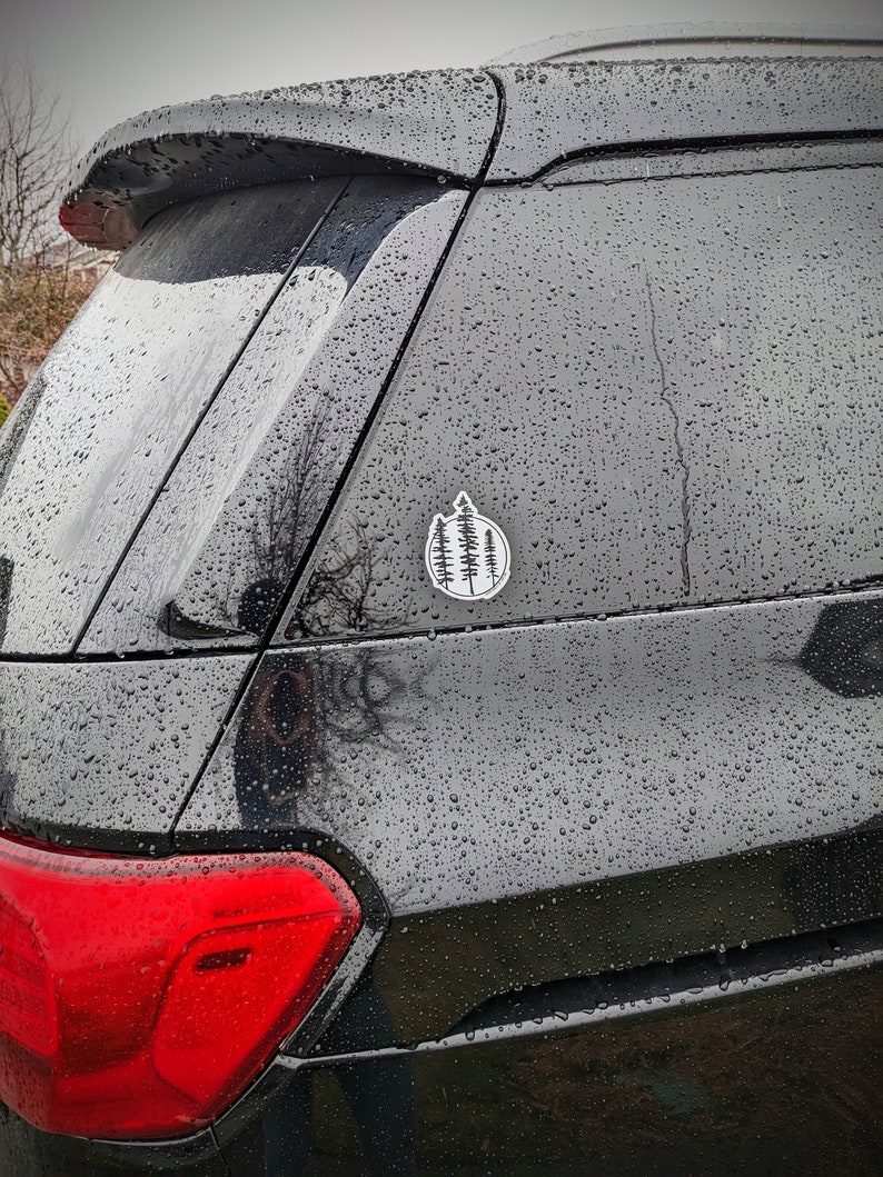 May include: A black car with a rear window covered in raindrops. A small white sticker with a black silhouette of a group of trees is on the rear window.