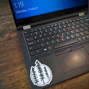 May include: A black laptop computer with a white sticker of three trees in a circle on the touchpad. The sticker has the text "MORE TREES PLEASE" written around the circle.