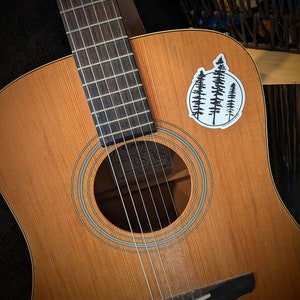 May include: A brown acoustic guitar with a black pickguard and a sticker of three pine trees on the body. The guitar has a soundhole with a white ring around it and a black bridge with six chrome tuning pegs.