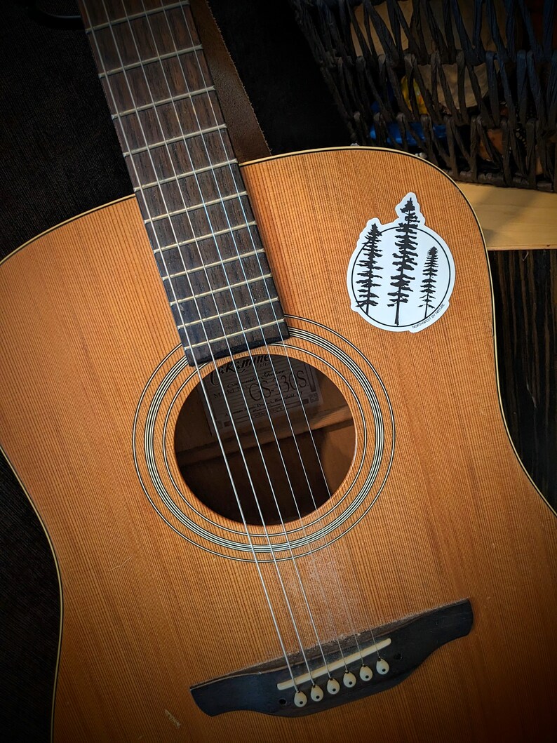 May include: A brown acoustic guitar with a black pickguard and a sticker of three pine trees on the body. The guitar has a soundhole with a white ring around it and a black bridge with six chrome tuning pegs.