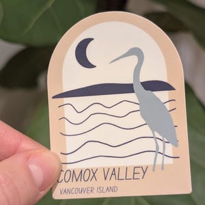 Peut inclure: Un autocollant avec une illustration stylisée d'un héron près de lignes ondulées, avec un croissant de lune. Le texte "Comox Valley Vancouver Island" est imprimé en bas.
