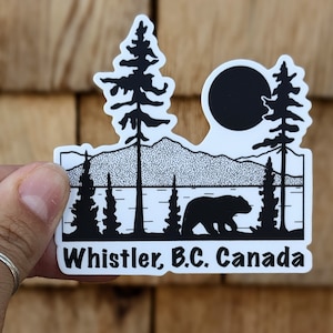 May include: Black and white sticker with a silhouette of a bear, trees, mountains, and a lake. The text "Whistler, B.C. Canada" is at the bottom of the sticker.