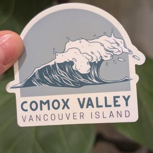 Peut inclure: Autocollant représentant une vague stylisée en bleu et blanc, avec le texte "COMOX VALLEY VANCOUVER ISLAND". L'autocollant a un fond bleu clair et une bordure blanche. Thème côtier.