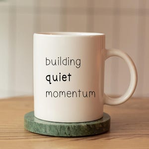 A white coffee mug with the text "building quiet momentum" printed on it, placed on a wooden table.