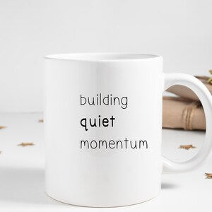 A white mug with the text "building quiet momentum" printed on it, placed on a white surface with scattered gold stars.