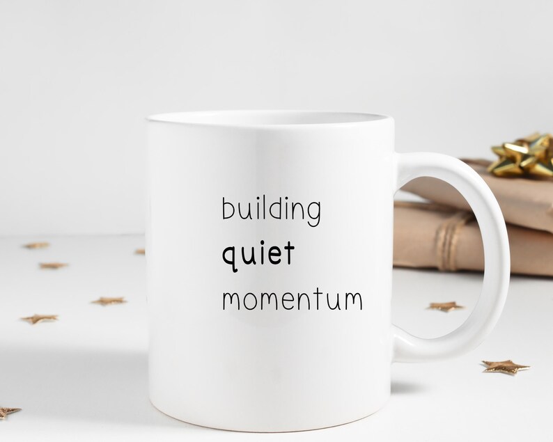 A white mug with the text "building quiet momentum" printed on it, placed on a white surface with scattered gold stars.