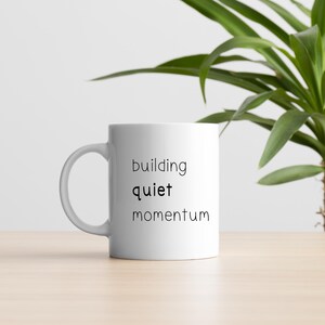 A white mug with the text "building quiet momentum" printed on it, placed on a wooden surface next to a potted plant.