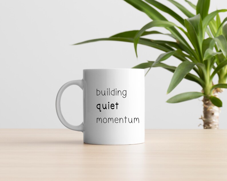 A white mug with the text "building quiet momentum" printed on it, placed on a wooden surface next to a potted plant.