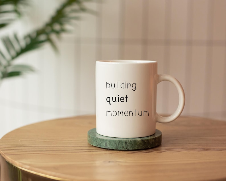 A white coffee mug with the text "building quiet momentum" printed on it, placed on a wooden table.