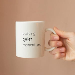 A hand holding a white mug with the text "building quiet momentum" printed on it.