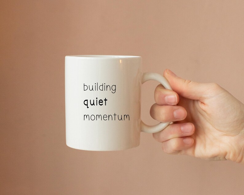 A hand holding a white mug with the text "building quiet momentum" printed on it.