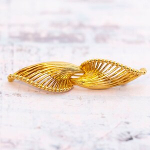 Puede incluir: Broche dorado con diseño de hojas. La broche presenta dos formas de hojas estilizadas conectadas en el centro. Cada hoja tiene una serie de líneas paralelas y un borde con cuentas. La broche está sobre un fondo claro.