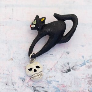May include: Black ceramic cat figurine with a white skull charm hanging from its tail. The cat is in a leaping pose with its head turned to the side. The skull charm has a painted face with black eyes and a toothy grin.
