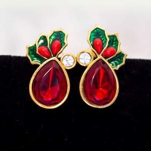 Può includere: Un paio di orecchini dorati con motivi a foglia di agrifoglio in smalto rosso e verde e accenti di cristallo rosso. Gli orecchini presentano cristalli rossi a forma di goccia.