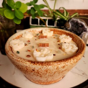 May include: A beige ceramic bowl with a brown glaze, filled with a white candle with embedded crystals and wooden sticks. The candle has a floral design on top.