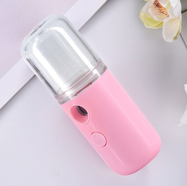 Pode incluir: Um pequeno pulverizador facial rosa de m&atilde;o com um reservat&oacute;rio de pl&aacute;stico transparente. O pulverizador tem um &uacute;nico bot&atilde;o no lado.