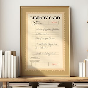 May include: A framed library card print with a vintage aesthetic. The card features the name "Olivia" and a list of book titles including "Anne of Green Gables" and "Pride and Prejudice". The frame is gold-toned, and the print is displayed on a wooden shelf with books.