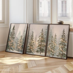 Könnte beinhalten: Drei gerahmte Kunstdrucke zeigen eine Winterwaldszene mit schneebedeckten Nadelbäumen. Das Kunstwerk hat eine weiche, gedämpfte Farbpalette mit warmen, funkelnden Lichtern, die eine ruhige Atmosphäre schaffen. Die Rahmen sind dunkelbraun.