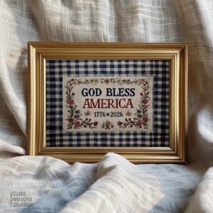 Può includere: Una cornice rettangolare color oro contenente un tessuto con motivo a quadretti blu e bianchi. Al centro, un pannello bianco riporta il testo "GOD BLESS AMERICA" in blu e rosso, con "1776 * 2026" sotto, incorniciato da ricami floreali.