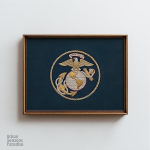 May include: A framed embroidery on a dark blue fabric background featuring the United States Marine Corps emblem. The design includes a gold eagle, globe, and anchor encircled by a rope border, with a banner reading Semper Fidelis.