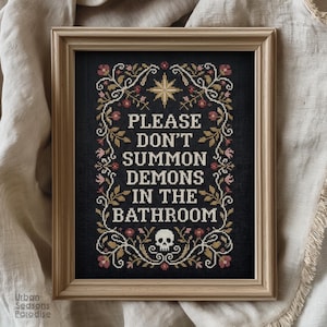 May include: A framed cross-stitch piece on black fabric featuring the white text PLEASE DON'T SUMMON DEMONS IN THE BATHROOM. The design is surrounded by a decorative border of gold vines, pink flowers, a gold star at the top, and a white skull at the bottom.