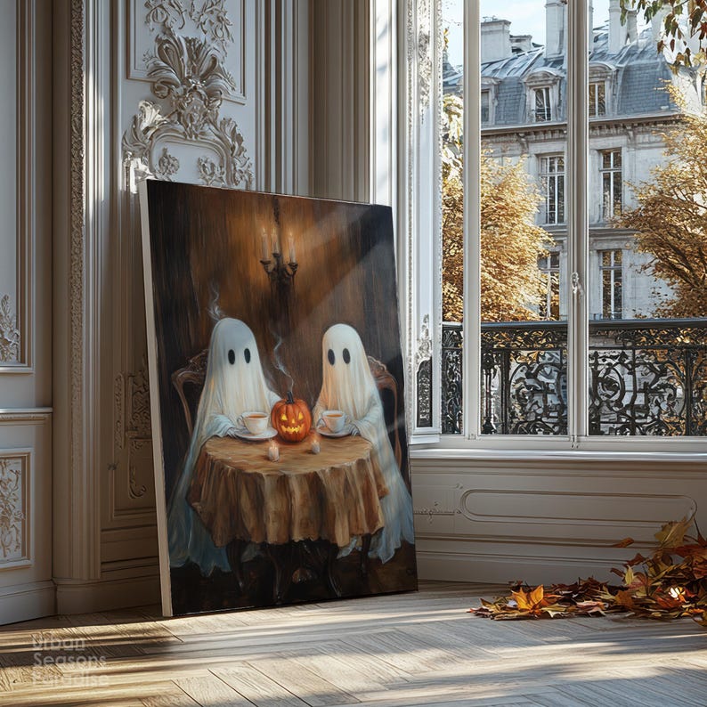 May include: A canvas print depicts two ghost figures seated at a table with a pumpkin and teacups. The artwork has a warm, inviting color palette with a vintage aesthetic. The print is leaning against a wall near a window with a view of a European building.