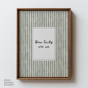May include: A framed print with a brown wooden frame. The background features vertical stripes in shades of green and white. A white rectangle in the centre displays the handwritten text "How lucky are we". The print is from Urban Seasons Paradise.