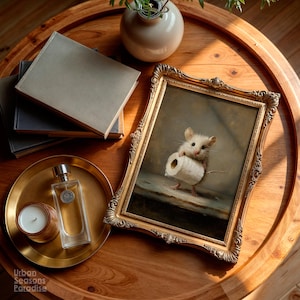 Op de afbeelding: Een ingelijste kunstdruk met een kleine muis die een rol toiletpapier vasthoudt, gepresenteerd in een sierlijke gouden lijst. Het kunstwerk ligt op een ronde houten tafel naast een stapel boeken, een glazen parfumflesje en een kaars.
