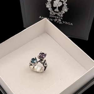 May include: A silver ring featuring two prong-set gemstones, one blue and one purple, with an open-work band design. The ring is presented in a white box with a black lid displaying the text JOYERIA ANGUS de la VEGA and a skull logo.