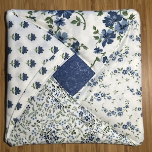 Puede incluir: Artículo de tela acolchada cuadrada con diseño de patchwork. El artículo presenta un cuadrado central de tela azul liso rodeado de secciones de tela blanca con estampados florales azules y ramitas florales azules más pequeñas. Los bordes están cuidadosamente cosidos.