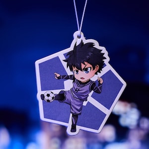 May include: A white air freshener with a blue hexagonal design. A cartoon character of a boy wearing a soccer uniform and kicking a soccer ball is printed on the air freshener.