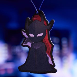 May include: A black and purple character with red hair, wearing a black cloak, is depicted on a car air freshener.
