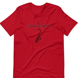 Puede incluir: Camiseta roja de manga corta con un gráfico de saxofón negro y el texto "I Still Believe".