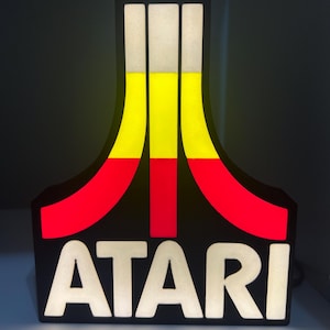 May include: Illuminated Atari logo sign. The sign features a black background with the word "ATARI" in white block letters. The iconic logo is composed of red, yellow, and white vertical stripes. The sign is lit, creating a retro aesthetic.