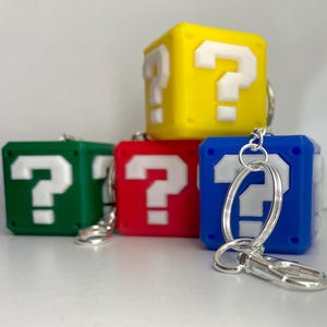 May include: Four colourful question mark block keychains. The keychains are made of soft, flexible plastic and feature a white question mark on each block. The blocks are yellow, green, red, and blue.