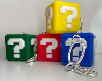 May include: Four colorful question mark block keychains. The keychains are made of soft, flexible plastic and feature a white question mark on each block. The blocks are yellow, green, red, and blue.