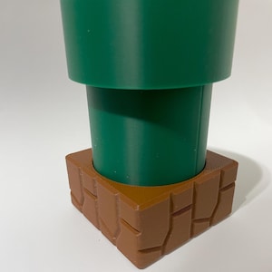 May include: A green and brown 3D printed model of a Super Mario Bros. pipe. The pipe is made of two pieces, a green top and a brown base. The base is textured to look like bricks.