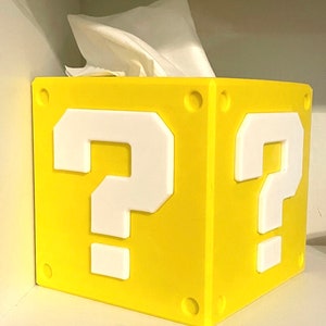 May include: A yellow, 3D printed tissue box shaped like a Super Mario Bros. question block with a white question mark on each side.