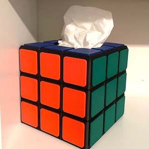 Puede incluir: Un porta pañuelos con forma de cubo de Rubik con cuadrados rojos, verdes y azules. La caja de pañuelos es blanca y es parcialmente visible en la abertura superior del porta pañuelos.