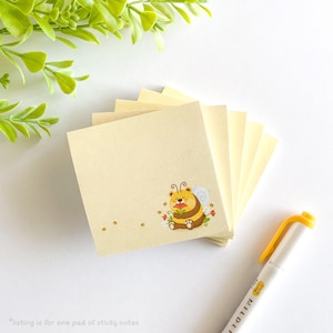 May include: A stack of yellow sticky notes with a cartoon bee illustration. The bee is wearing a flower and has a happy expression. The notes are perfect for adding reminders, notes, or decorations to your workspace.