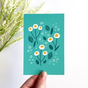 May include: A teal blue card with a white background featuring a stylized illustration of white flowers with yellow centers and green leaves. The flowers are arranged in a cluster and are surrounded by small white circles.