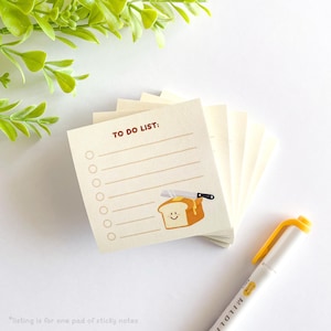 May include: A stack of square sticky notes with a to-do list design. The notes are cream colored with brown lines and a cartoon illustration of a slice of bread with butter and a knife. The text "TO DO LIST:" is printed in brown at the top of the notes.