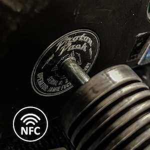 May include: Close-up of a black metal object with a circular sticker that reads "Proton Pack" and "SERIAL # 2 OPERATOR: JAMIE FUCA". The object has a silver metal rod extending from the center of the sticker. A white circle with a wifi symbol and the text "NFC" is in the lower left corner of the image.