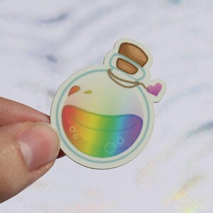 May include: A clear glass bottle filled with rainbow-coloured liquid, featuring a cork stopper and a small pink heart charm. The bottle is set against a white background.