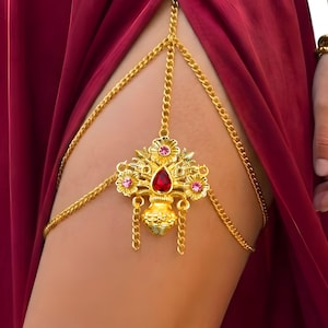May include: A gold-toned metal thigh chain featuring a central floral-inspired ornament. The centrepiece includes a prominent red teardrop-shaped stone, smaller pink circular stones, and dangling chain accents, all connected by delicate gold chains.