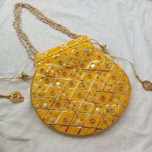 May include: A yellow round purse with a gold chain strap and gold embellishments. The purse is decorated with a geometric pattern of gold and silver.
