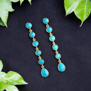 May include: A pair of gold-toned earrings with turquoise-colored stones. Each earring features a series of round stones and a teardrop-shaped stone at the bottom. The earrings are displayed on a dark background with green leaves.