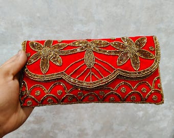 May include: A red clutch with gold beaded floral and geometric designs. The flap closure and edges are also adorned with gold beads. The clutch is held in a hand, showcasing the intricate details of the beaded embellishments.