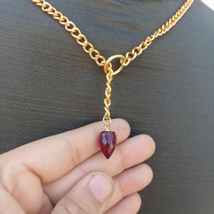 Puede incluir: Un collar de cadena dorada con un colgante de piedra roja. El colgante es facetado y tiene una punta puntiaguda.